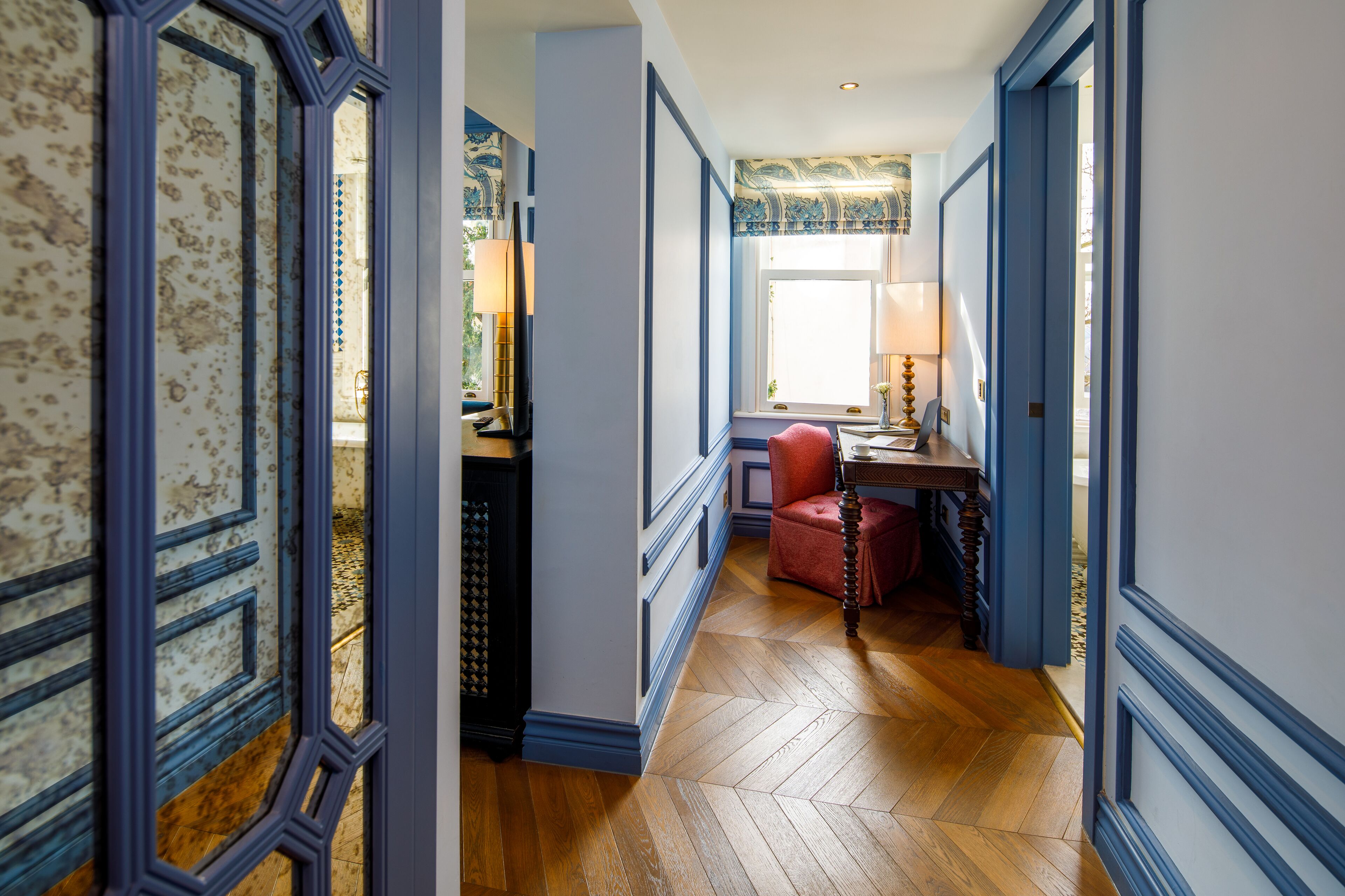 A hallway with wooden flooring, a red chair, and a desk with a lamp.