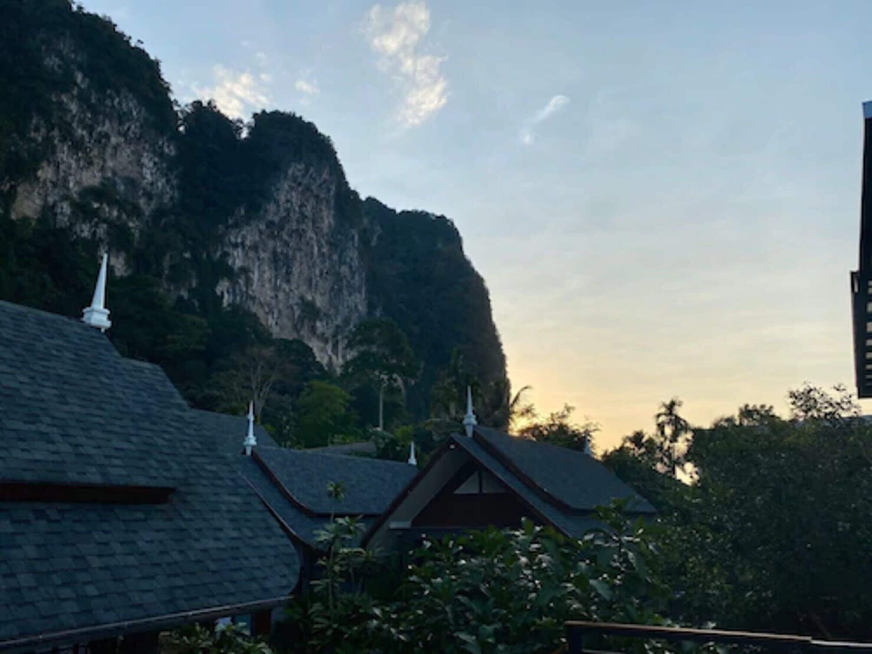 A traditional building with steeply pitched roofs and white decorative elements, surrounded by lush greenery and a large cliff.