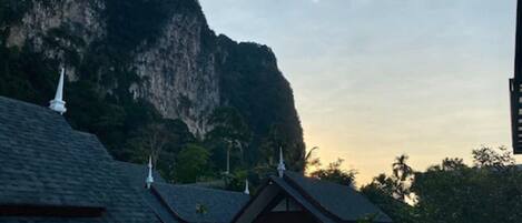 A traditional building with steeply pitched roofs and white decorative elements, surrounded by lush greenery and a large cliff.