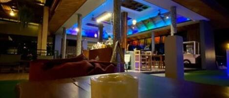 Un espacio interior con una mesa de madera y un vaso de una bebida amarilla. Hay una zona de estar con un sofá rojo y una persona al fondo.
