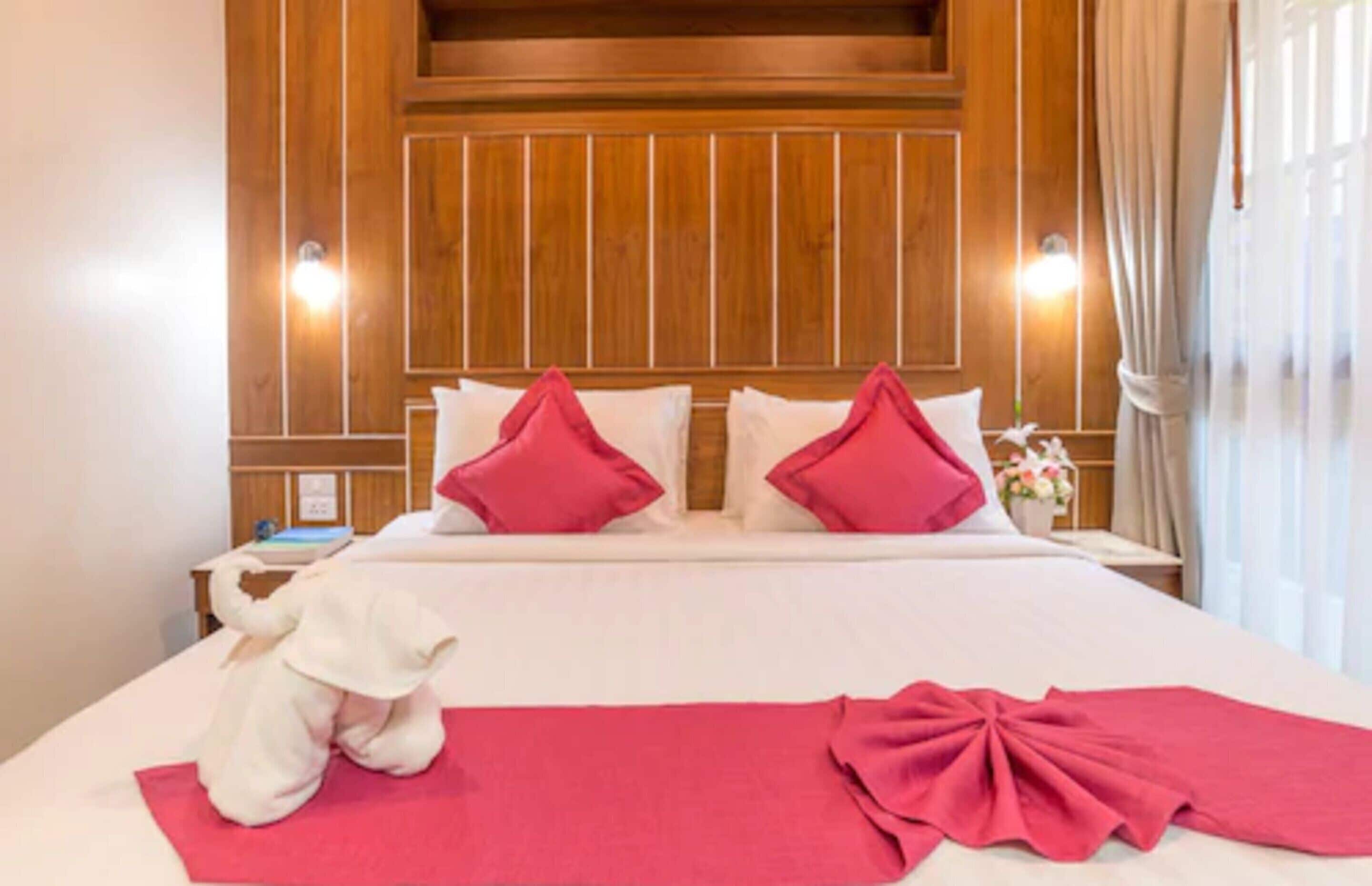 A hotel room with a wooden headboard, two pillows, a bed with a red blanket, a white stuffed animal, and a nightstand with a lamp.