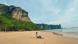 A beach with a person sitting on the sand, cliffs in the background, and a clear sky. - The Scene Cliff View Villas (Krabi)