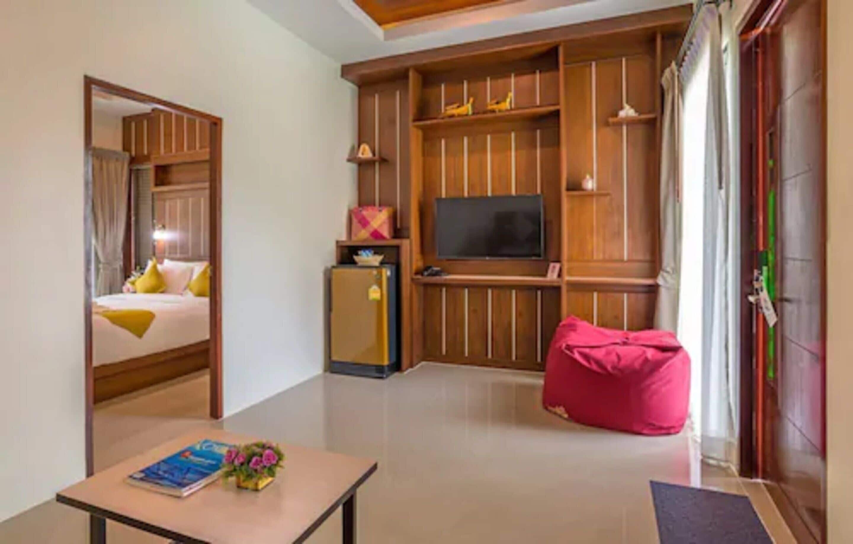 A hotel room with a bed, a television, a small refrigerator, a pink bean bag, and a table with a book and flowers.