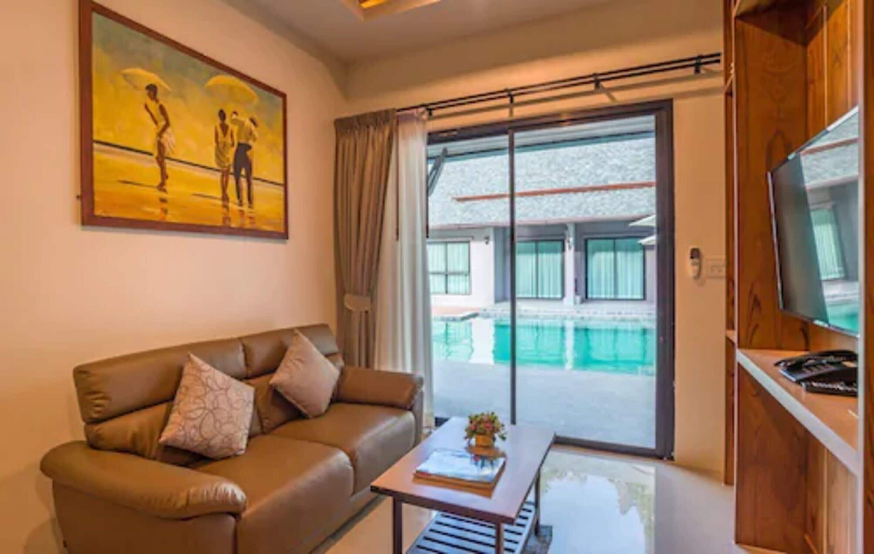 Una sala de estar con un sofá de cuero marrón, una mesa de centro con un libro y flores, un cuadro en la pared y un ventanal corredizo que da a una zona de piscina.