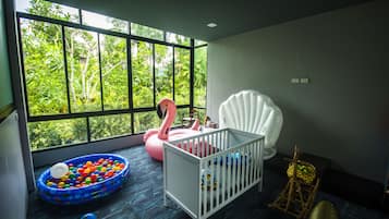 Una habitación con una cuna, una mecedora, un ventanal con vistas a follaje y una zona de juegos con piscina de pelotas y juguetes inflables.