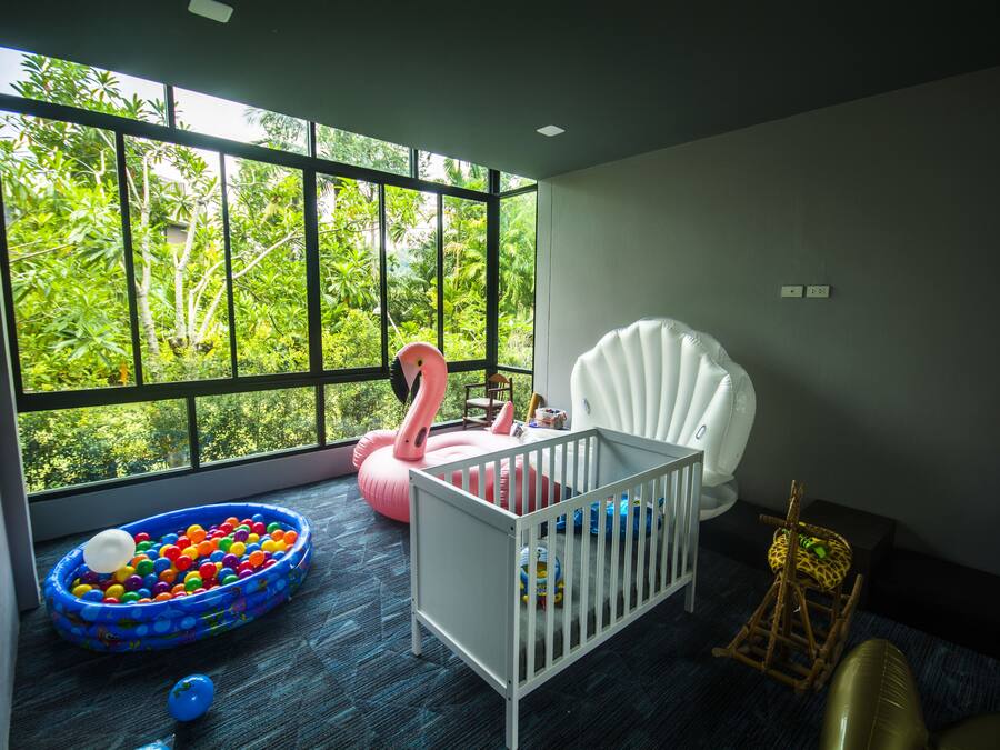 Una habitación con una cuna, una mecedora, un ventanal con vistas a follaje y una zona de juegos con piscina de pelotas y juguetes inflables.
