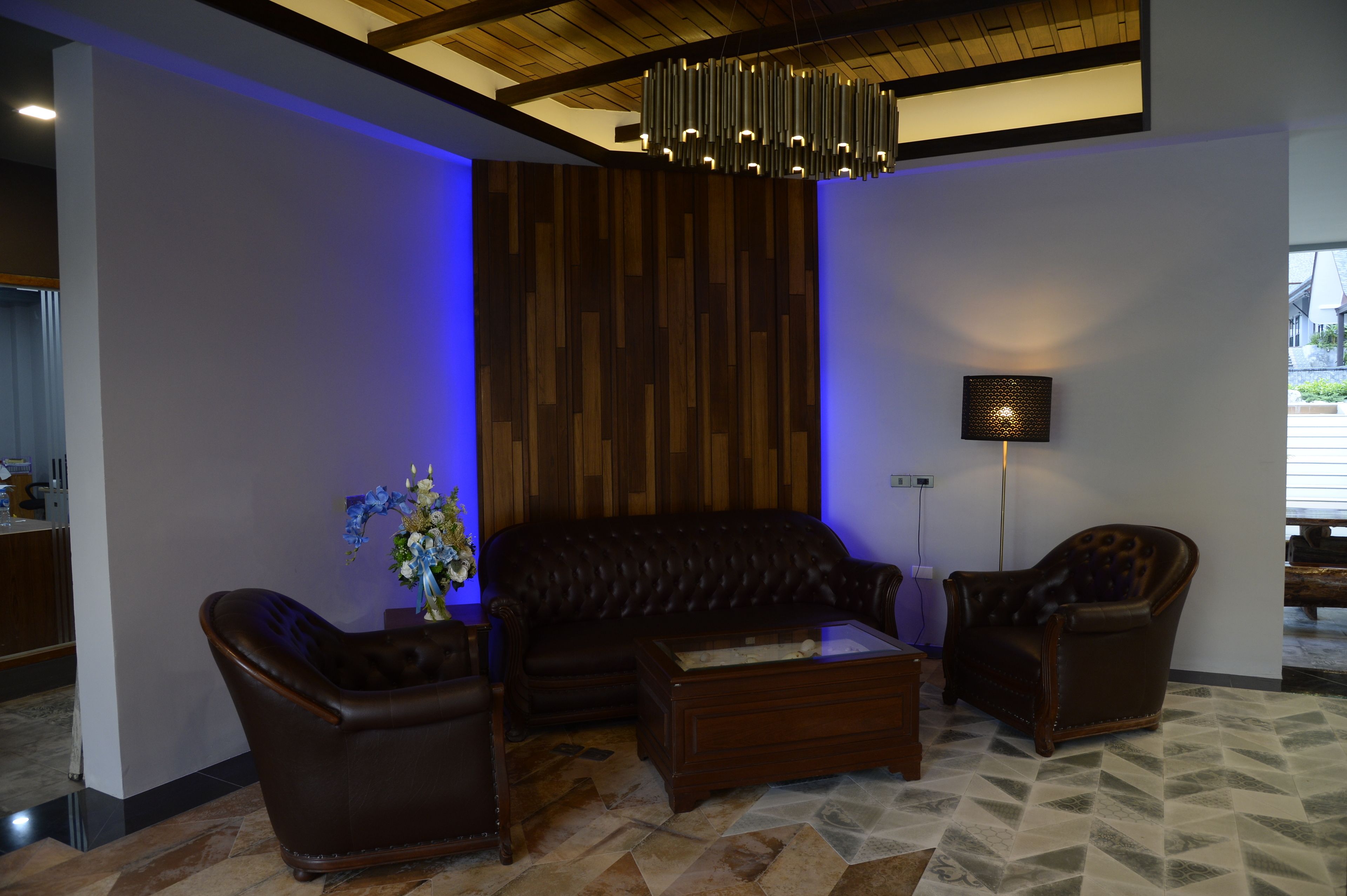 A modern living room with a leather sofa, two armchairs, and a coffee table. There is a wooden accent wall, a floor lamp, and a vase with flowers.