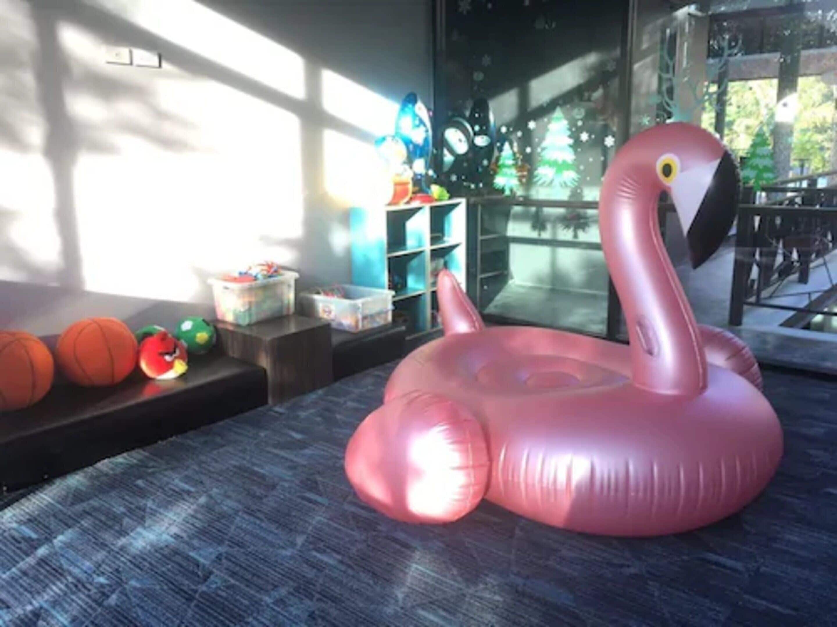 A room with a large pink inflatable flamingo, a window with blinds, a shelf with toys, and a small table.