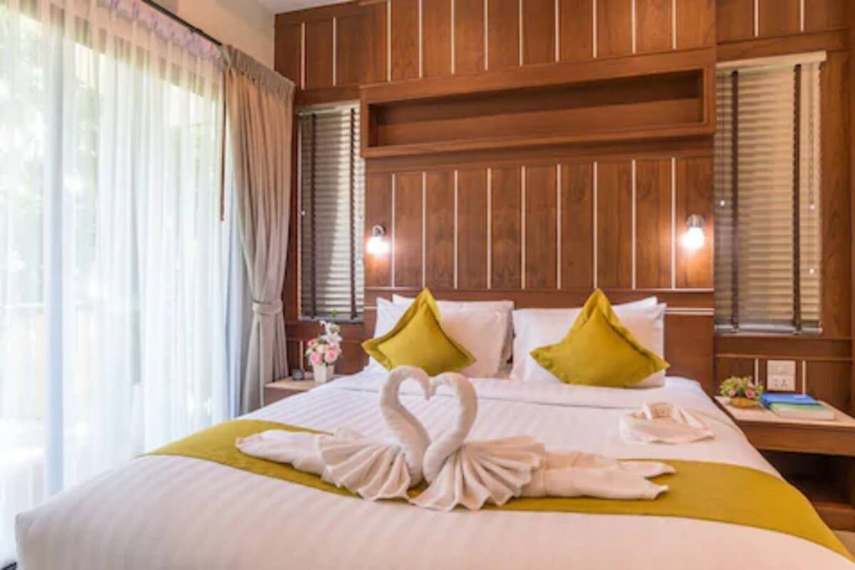A hotel room with a wooden panel wall, a bed with white bedding and yellow pillows, and a swan-shaped towel arrangement.