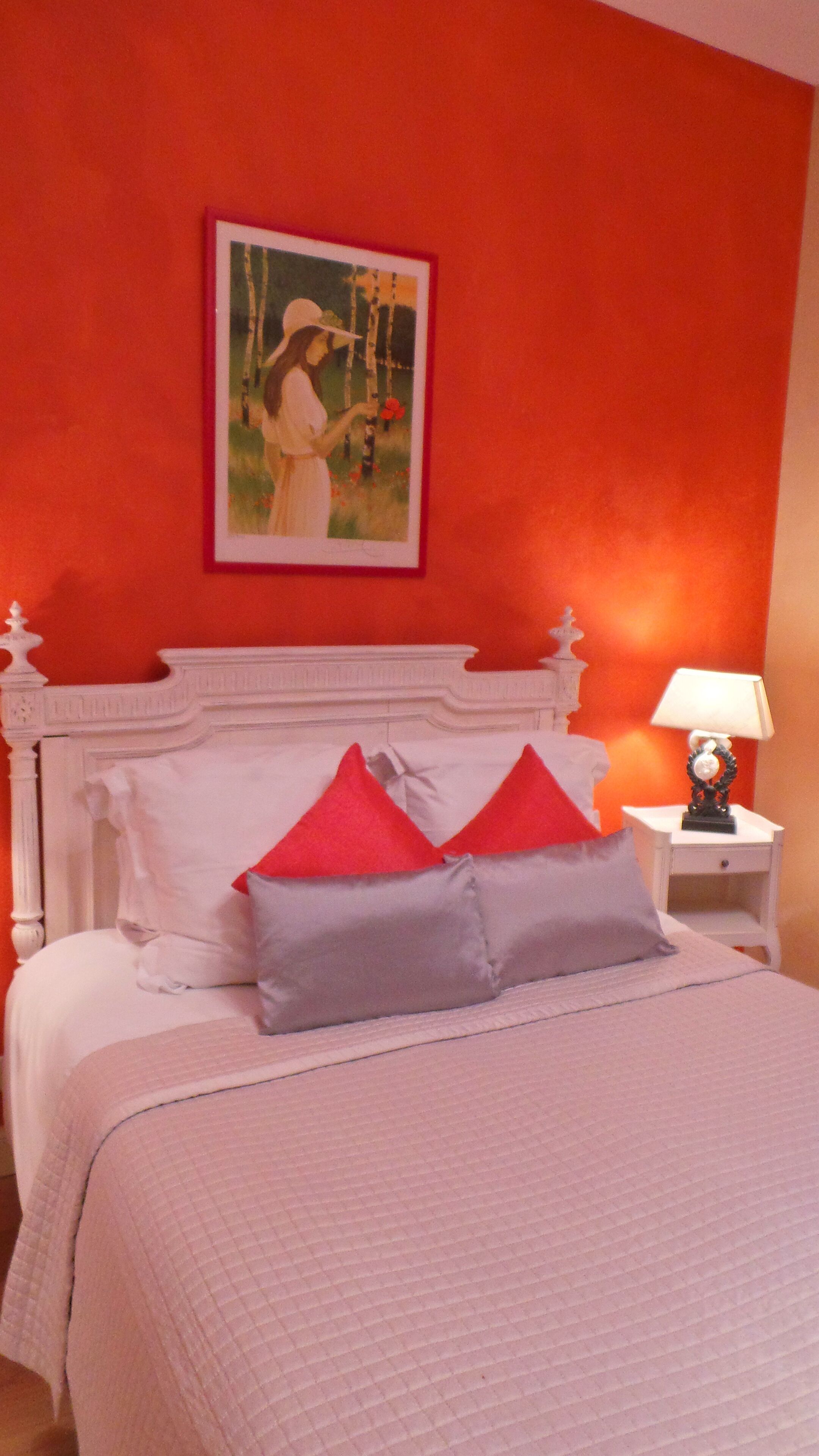 Une chambre avec un lit blanc, des oreillers rouges et gris, une table de chevet blanche avec une lampe, et un tableau représentant une femme accroché au mur.
