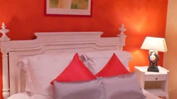 Une chambre avec un lit blanc, des oreillers rouges et gris, une table de chevet blanche avec une lampe, et un tableau représentant une femme accroché au mur.