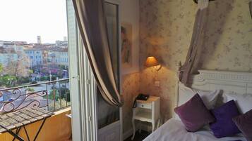 Habitación de hotel con balcón, cama con almohadas, mesita de noche con lámpara y vista a edificios.
