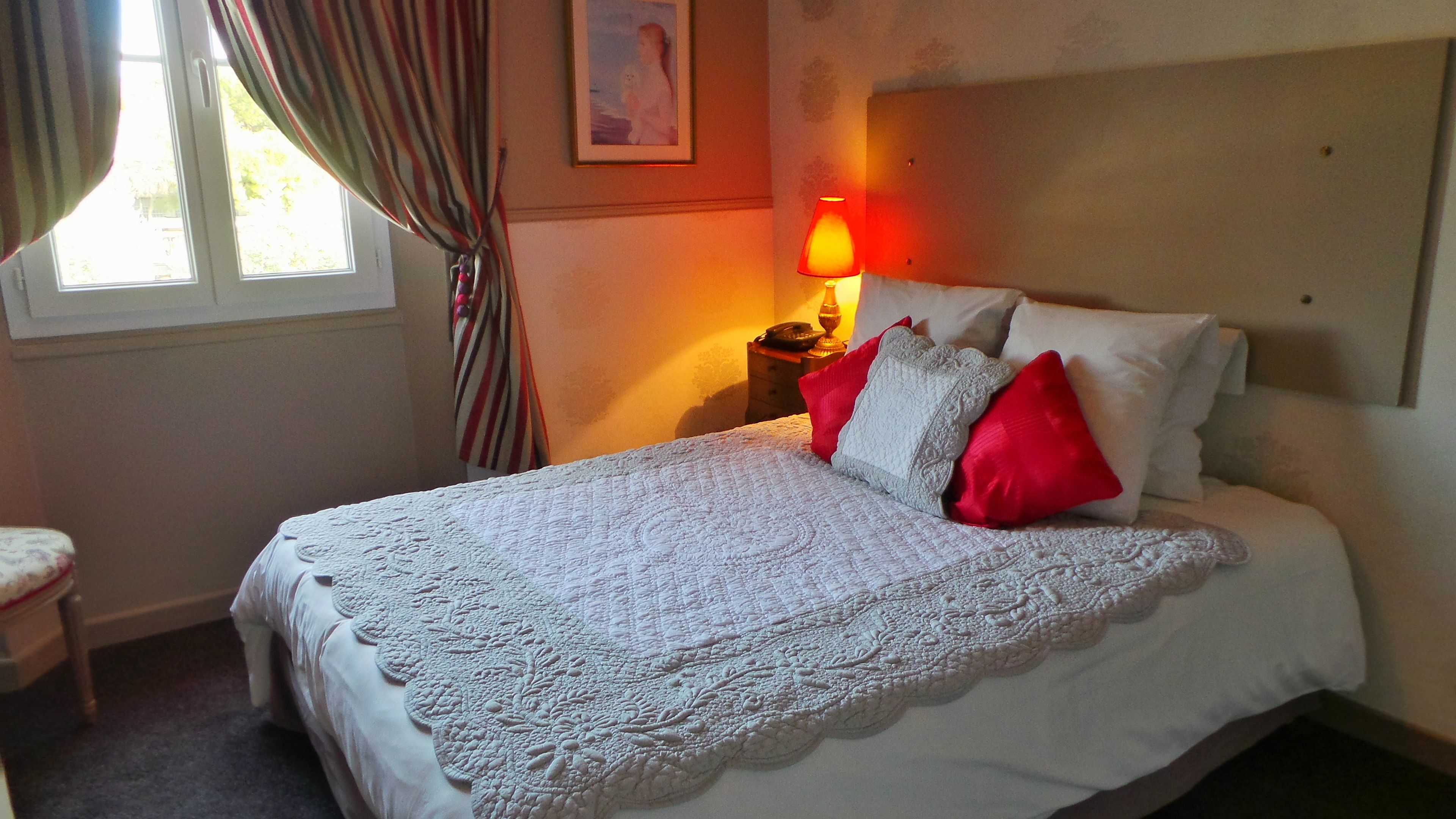 A neatly made bed with a white lace bedspread and red pillows, a bedside lamp, and a framed picture on the wall.
