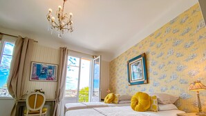 A bedroom with two beds, a chandelier, a chair, and a window with curtains. - Hotel Le Relais Du Postillon (Antibes)