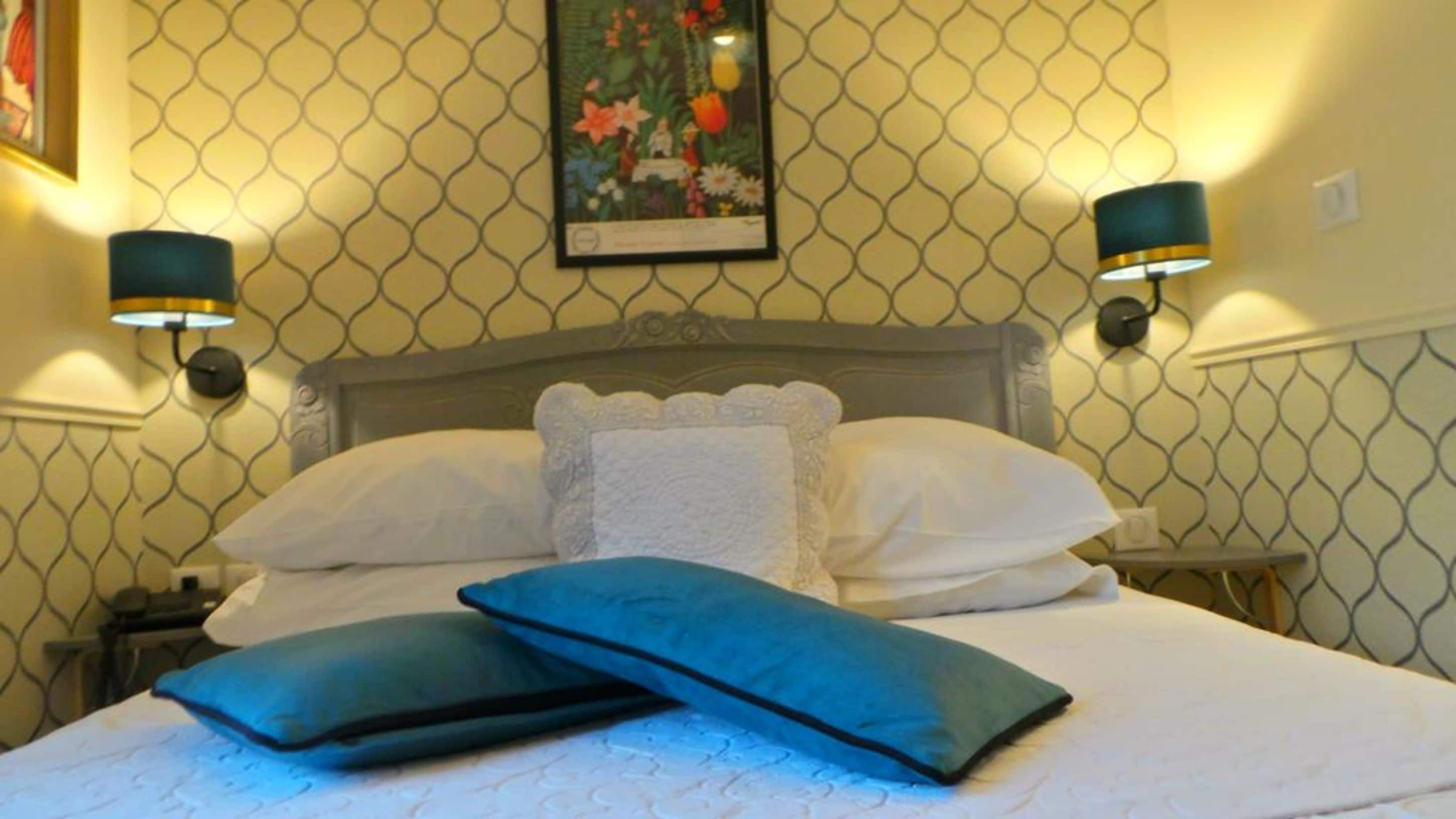 Ein ordentlich bezogenes Bett mit zwei blauen Kissen, einem dekorativen Kopfteil und zwei Wandleuchten.