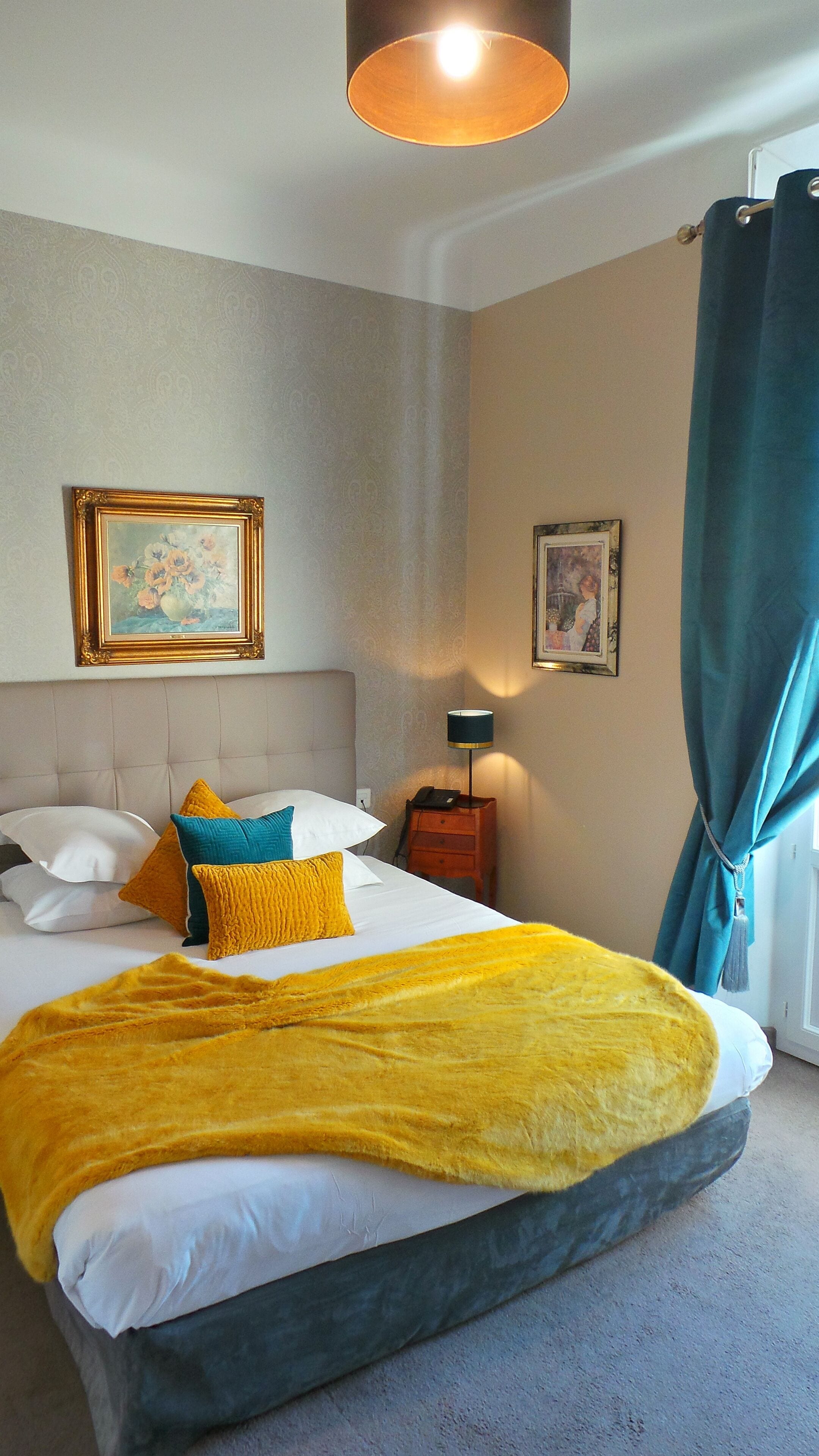 Ein Hotelzimmer mit einem Bett, zwei gerahmten Bildern, einem Nachttisch, einer Lampe und einem Vorhang.