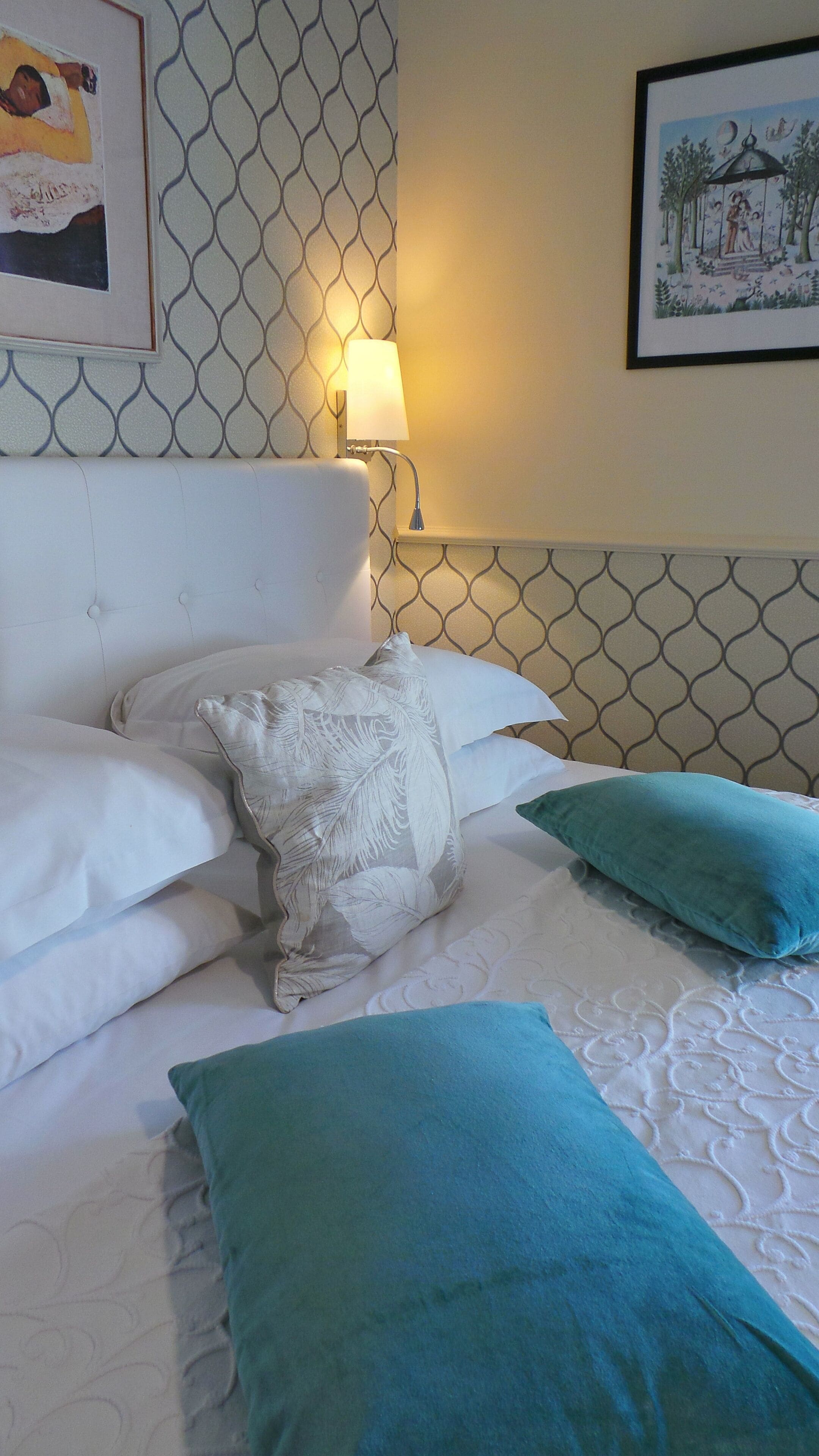 A neatly made bed with white and teal pillows, a wall-mounted lamp, and a patterned wallpaper.