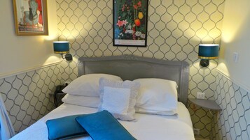 Ein ordentlich bezogenes Bett mit weißen Leinen und einem dekorativen Kopfkissen, zwei Wandlampen und ein gerahmtes Blumenbild über dem Bett.