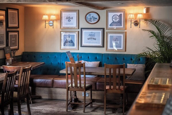 A cozy bar area with a blue cushioned bench, wooden tables, and chairs. There are framed pictures on the wall and wall-mounted lamps. - The Dog & Partridge by Greene King Inns (Bury St Edmunds)