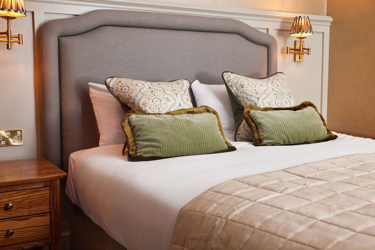 Een opgemaakt bed met een grijs beklede hoofdbord, witte lakens en decoratieve kussens.