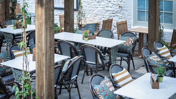 Café al aire libre con mesas y sillas, rodeado por un muro de piedra y postes de madera.