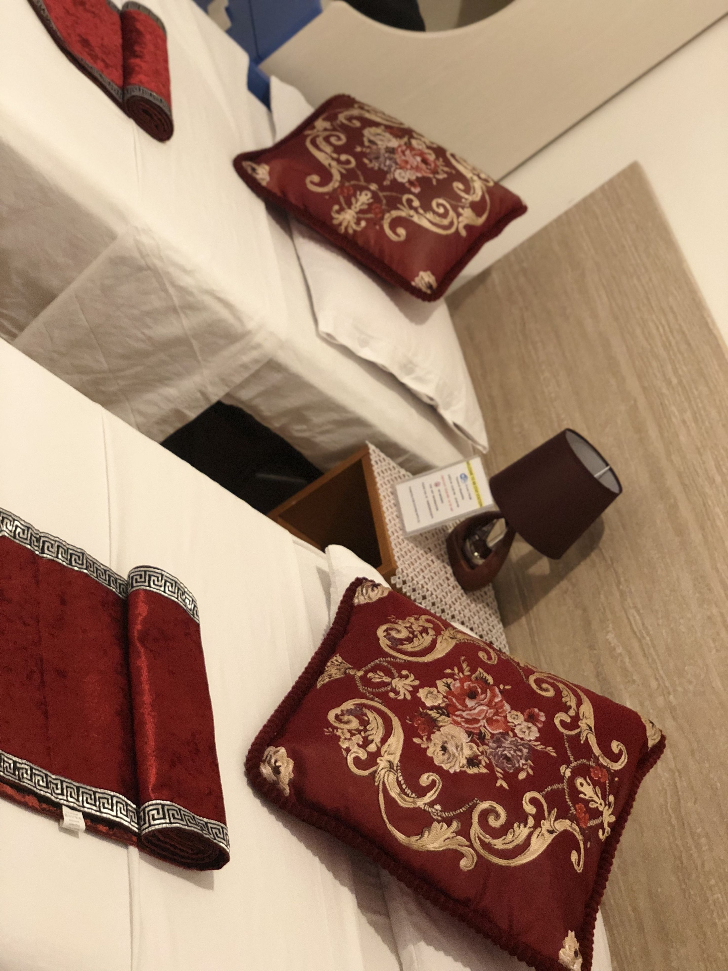 A hotel room with neatly arranged beds, each with a red pillow and a white bedspread. There is a small table with a lamp and a sign, likely indicating room amenities or information.
