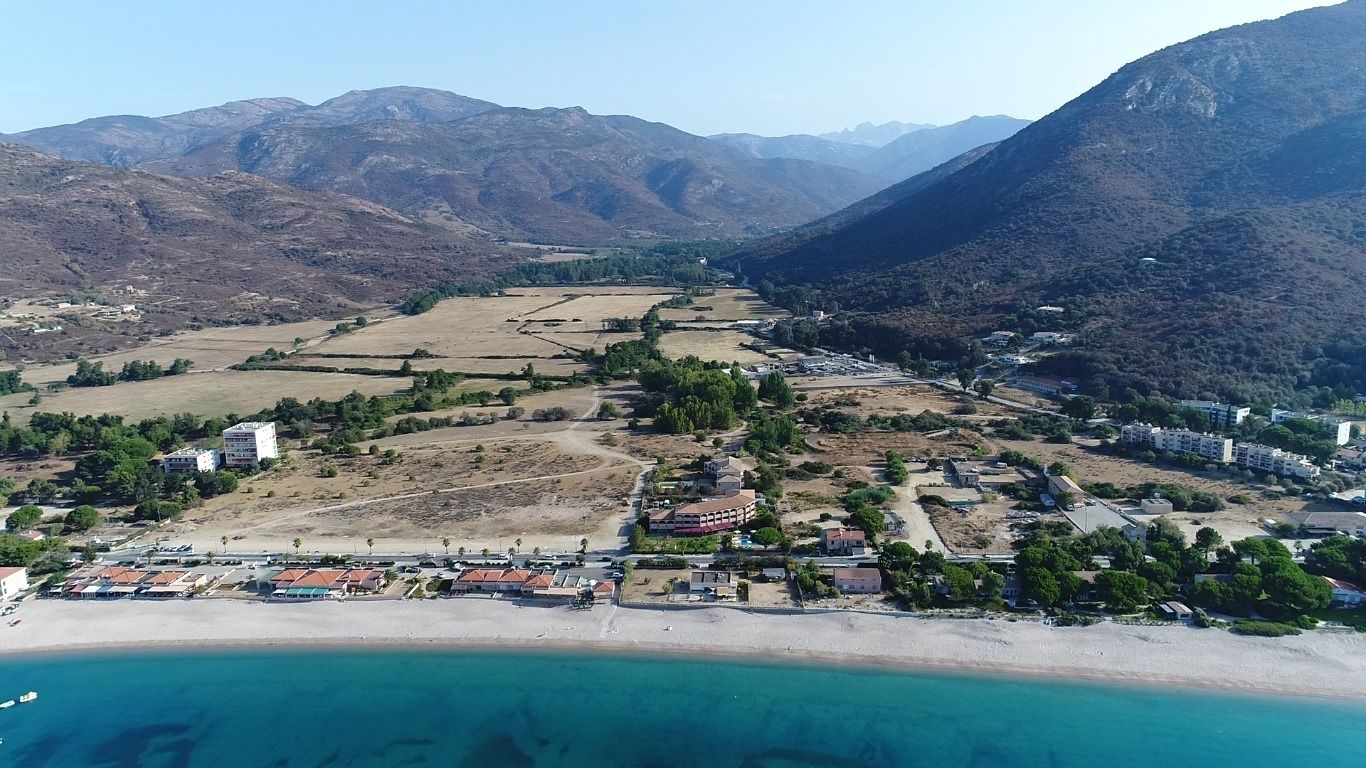 An aerial view of a coastal town with a beach, residential buildings, and mountainous terrain in the background.