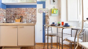 Une cuisine avec un mur recouvert de briques, un évier et une fenêtre. On y trouve des tabourets et une table sur laquelle sont posés divers objets.