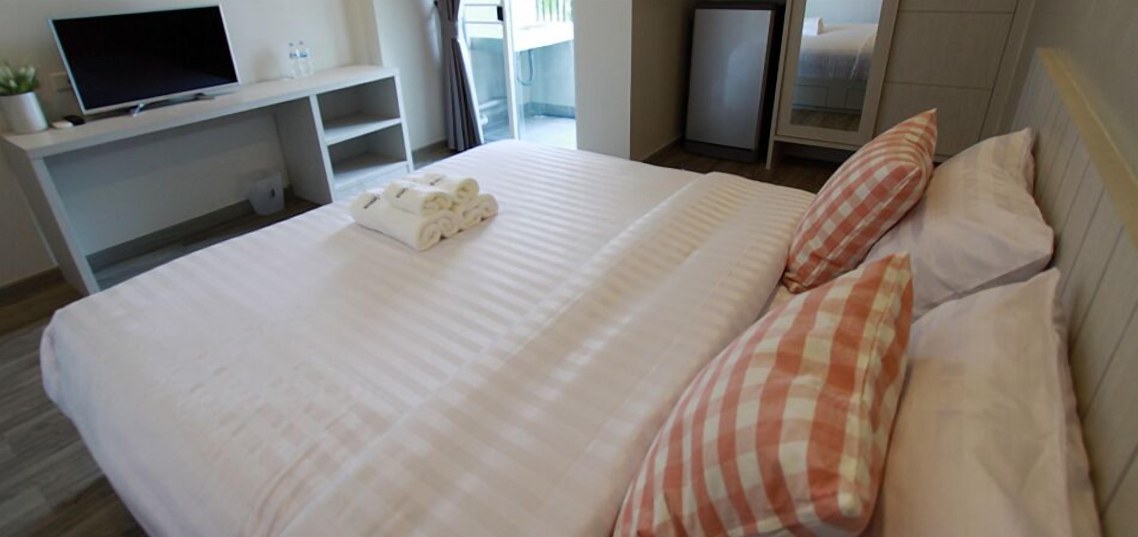 A hotel room with a neatly made bed, a flat-screen TV on a stand, a small desk with a computer, and a window with curtains.