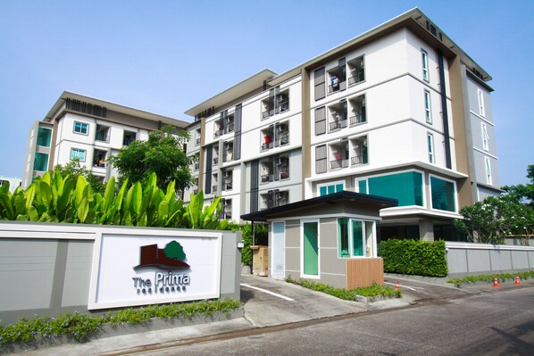 Un immeuble résidentiel à plusieurs étages, doté de balcons, d’une enseigne indiquant « The Prima », et d’un petit espace d’entrée avec un portail.