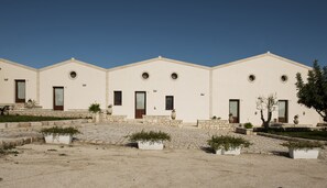 A row of beige buildings with circular windows and doors, stone steps leading to the entrance, and potted plants in the foreground. - I 5 sensi (Modica)