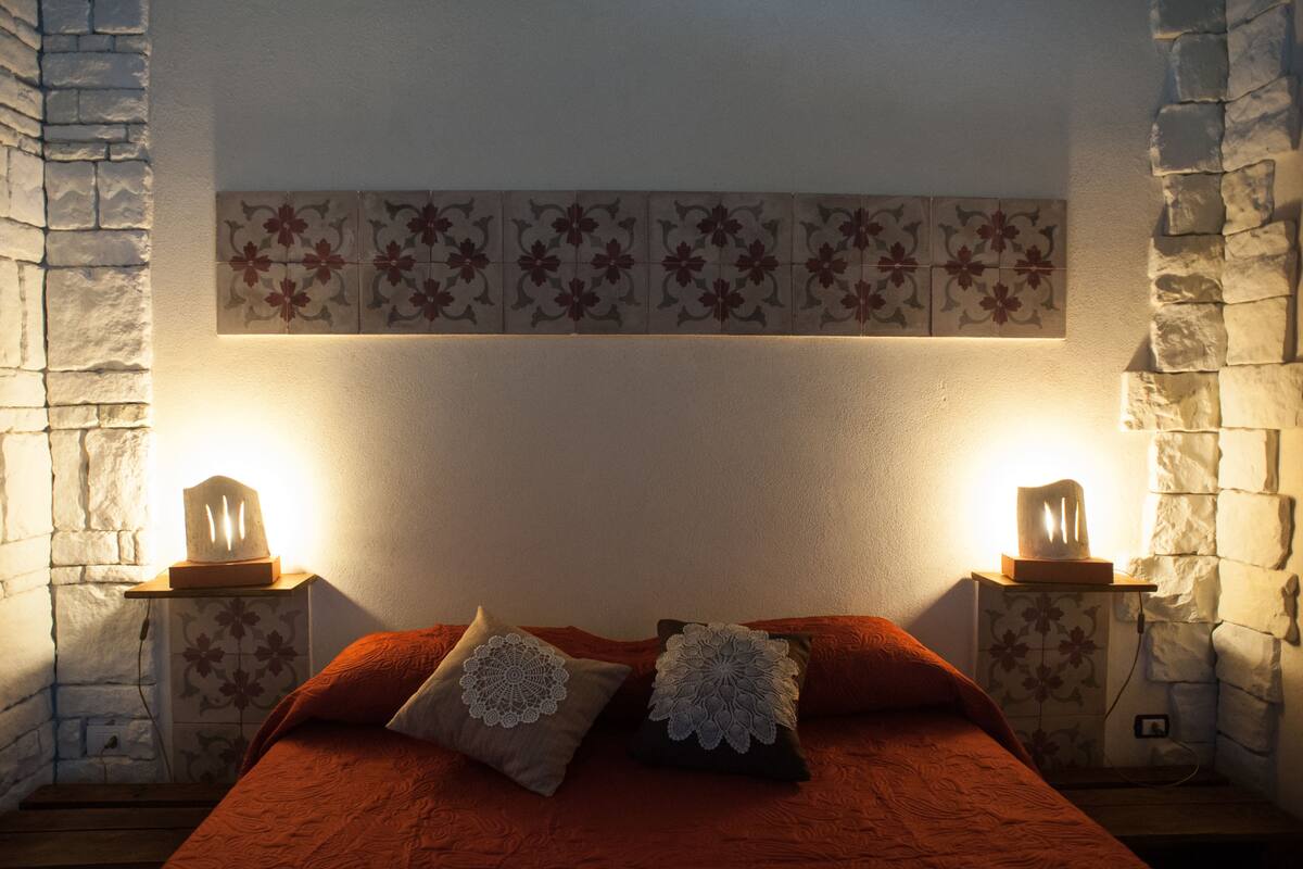 Une chambre avec un mur en pierre, un lit recouvert d’une couverture rouge et deux lampes de chevet.