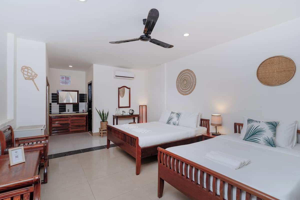 A hotel room with two wooden beds, a ceiling fan, a desk with a chair, a small table with a lamp, and a wall-mounted air conditioner.