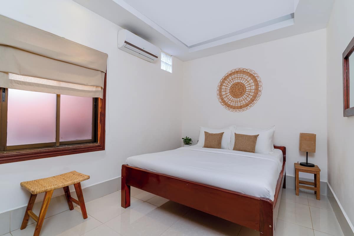 A neatly arranged bedroom with a large bed, a small stool, a wall-mounted lamp, and a decorative wall piece.