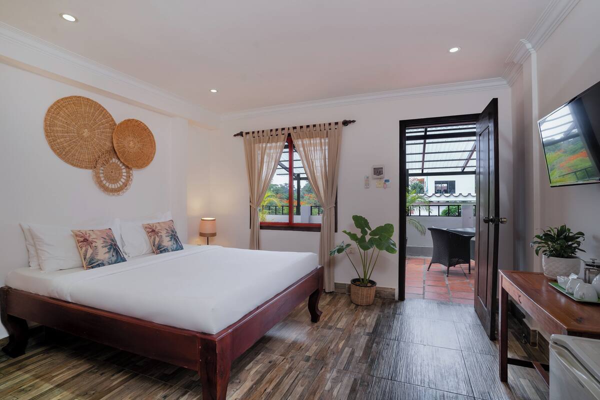 A bedroom with a large bed, a potted plant, a desk, and a view through an open door.
