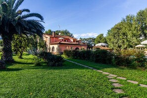 Una villa roja con un sendero en el jardín que lleva a la entrada, rodeada de vegetación exuberante y una palmera.
