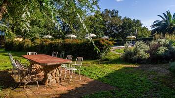 Een terras buiten met een stenen tafel en metalen stoelen, omringd door weelderige begroeiing en parasols.