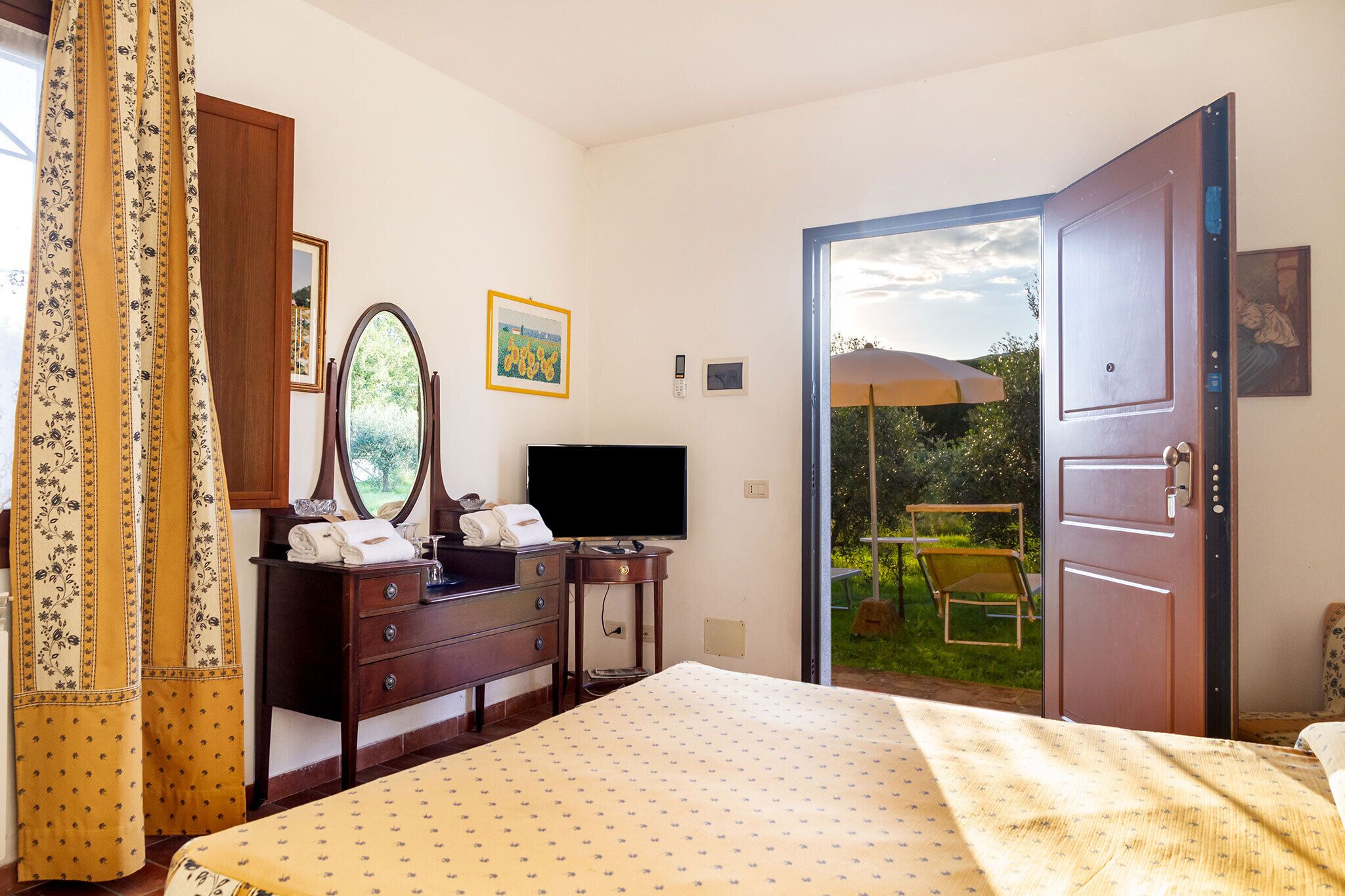 Une chambre à coucher comprenant un lit, une commode, un téléviseur et une porte donnant sur un balcon équipé d’une chaise et d’un parasol.