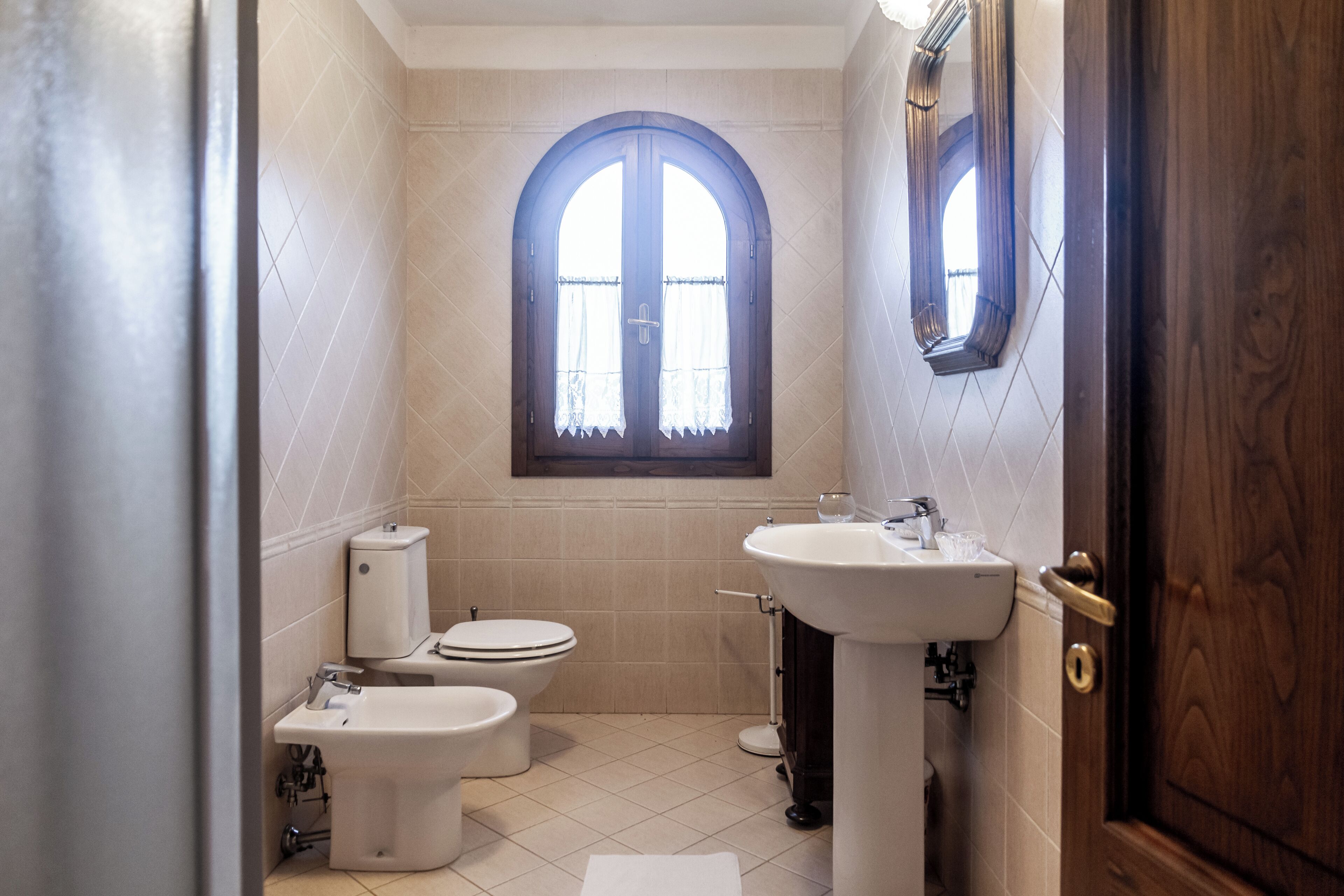 Un baño con inodoro, bidé y lavamanos, una ventana con cortinas y una puerta de madera.