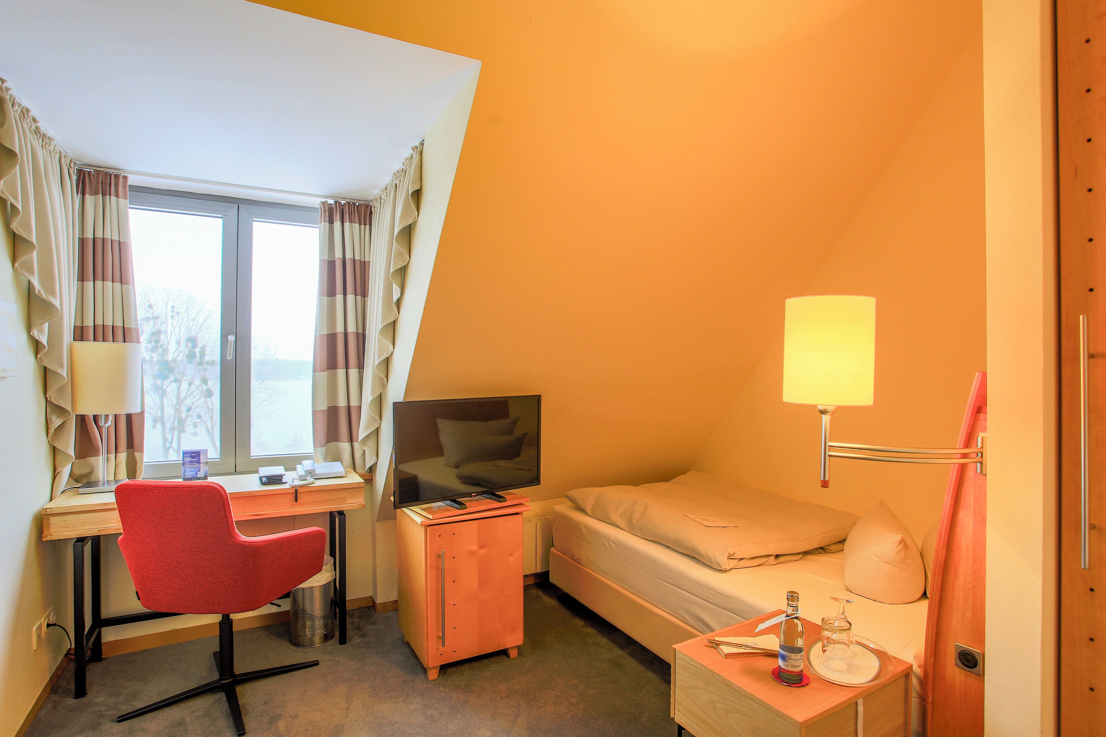 Habitación de hotel con escritorio, silla, cama, televisor y mesita de noche con lámpara.