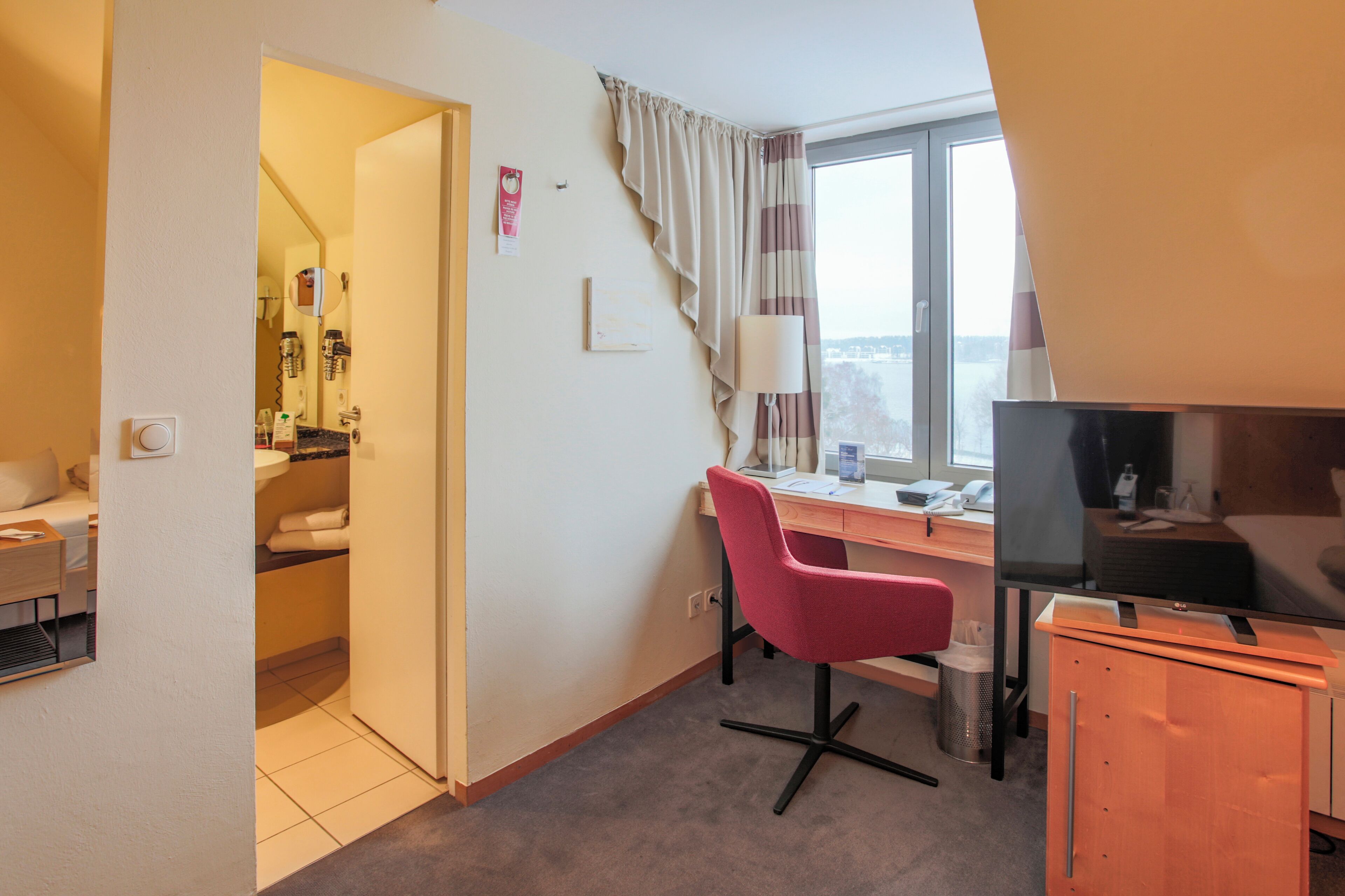 A hotel room with a desk, chair, TV, and a view of the city.