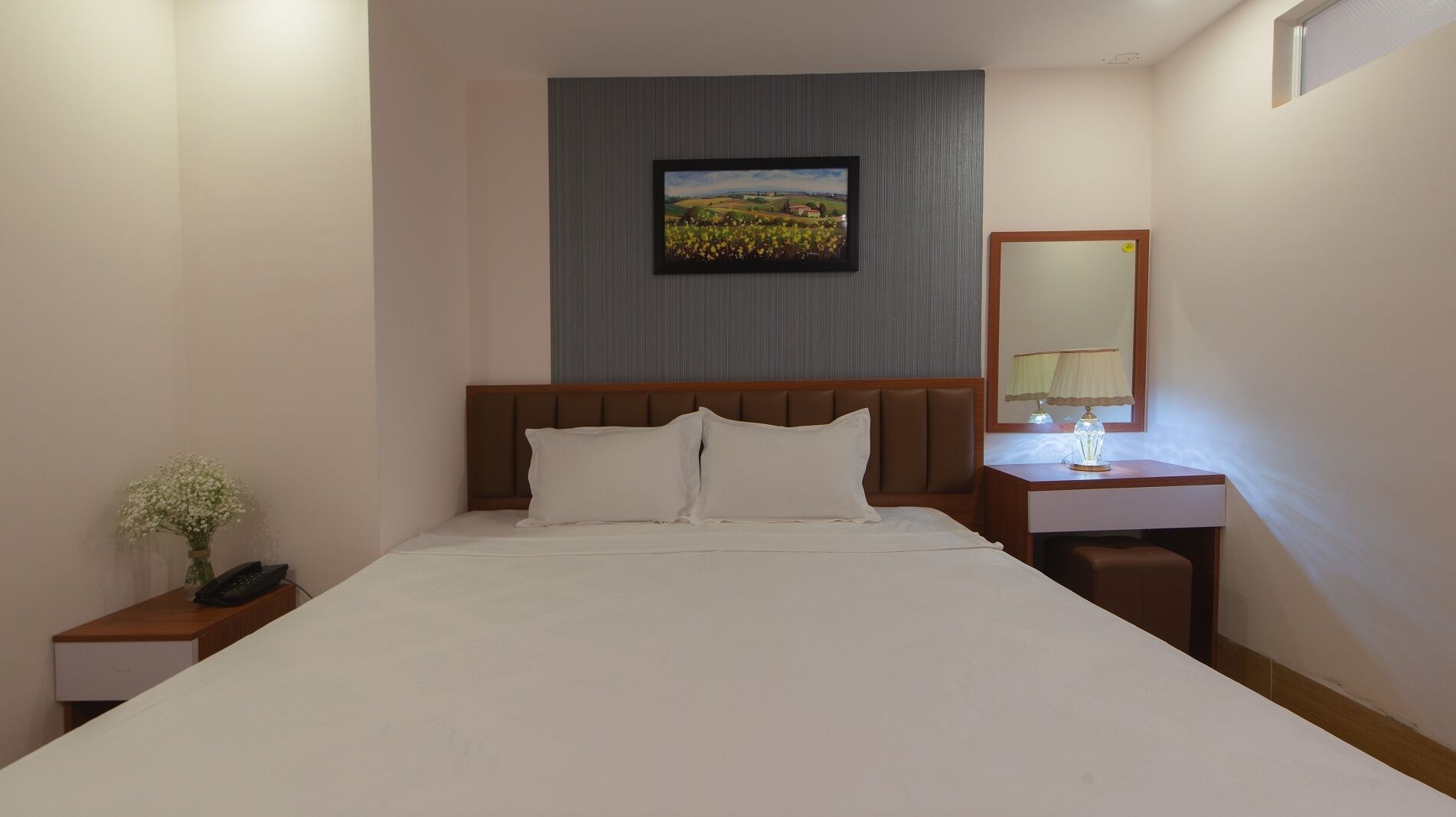 A hotel room with a bed, a nightstand with a phone, a bedside table with a lamp, and a painting on the wall.