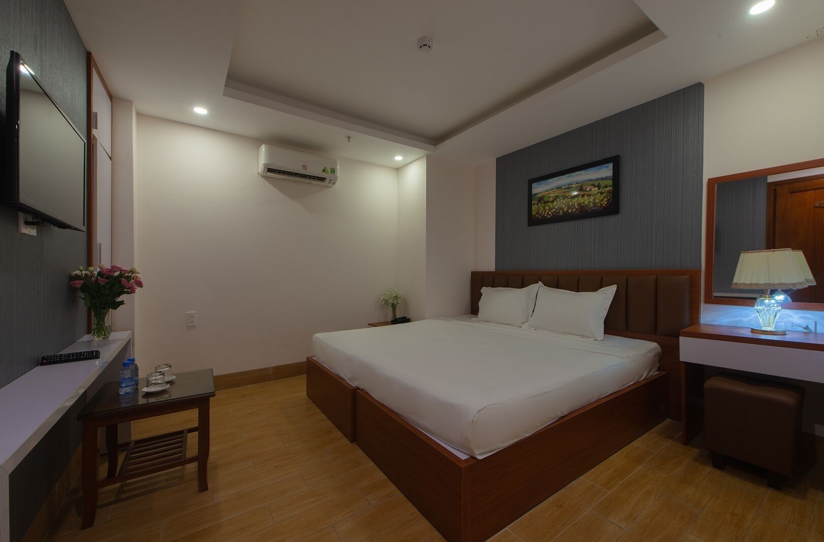 A hotel room with a bed, bedside table, lamp, air conditioning unit, and a painting on the wall.
