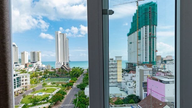 A cityscape through a window with a view of buildings, roads, and a construction crane. - Canary Nha Trang Hotel (Bac Nha Trang)