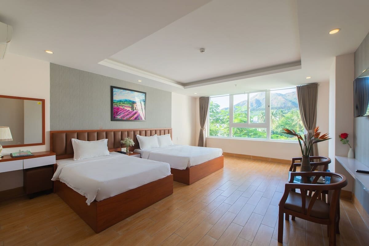 Une chambre d’hôtel avec deux lits, un bureau, une chaise, une fenêtre donnant sur un espace verdoyant et un tableau au mur.