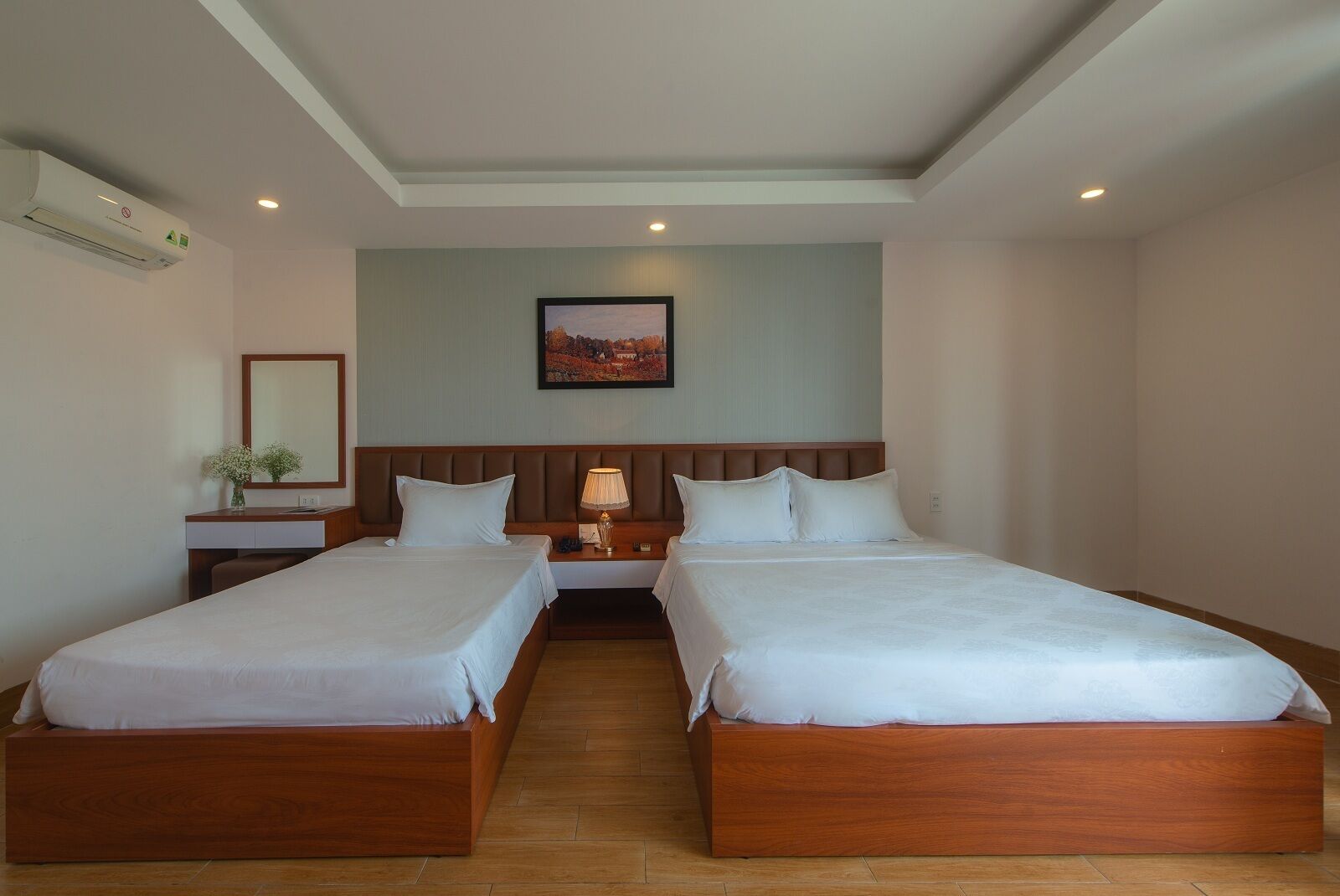 A hotel room with two single beds, a nightstand with a lamp, a wall-mounted air conditioner, and a framed picture above the beds.