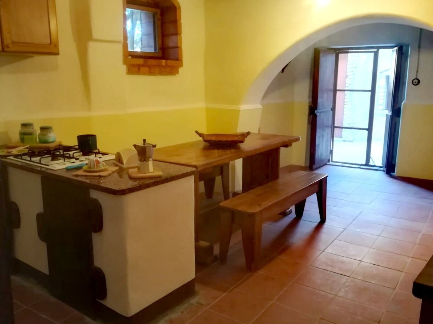 Een keuken met een gasfornuis, een houten tafel, een bankje en een klein raam.