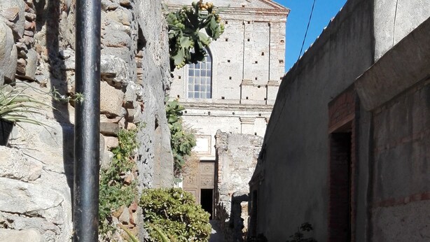A narrow street with a stone building featuring a cross on top, potted plants, and a cactus in the foreground. - Case Rurali Pentedattilo (Melito di Porto Salvo)