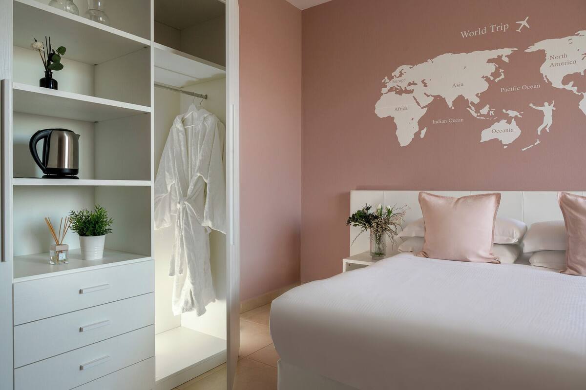 A bedroom with a white bed, a world map on the wall, a white wardrobe, and a nightstand with a lamp.
