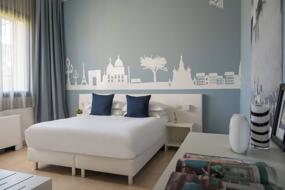 A modern hotel room with a large bed, a nightstand, a bedside table, and a cityscape wall mural.